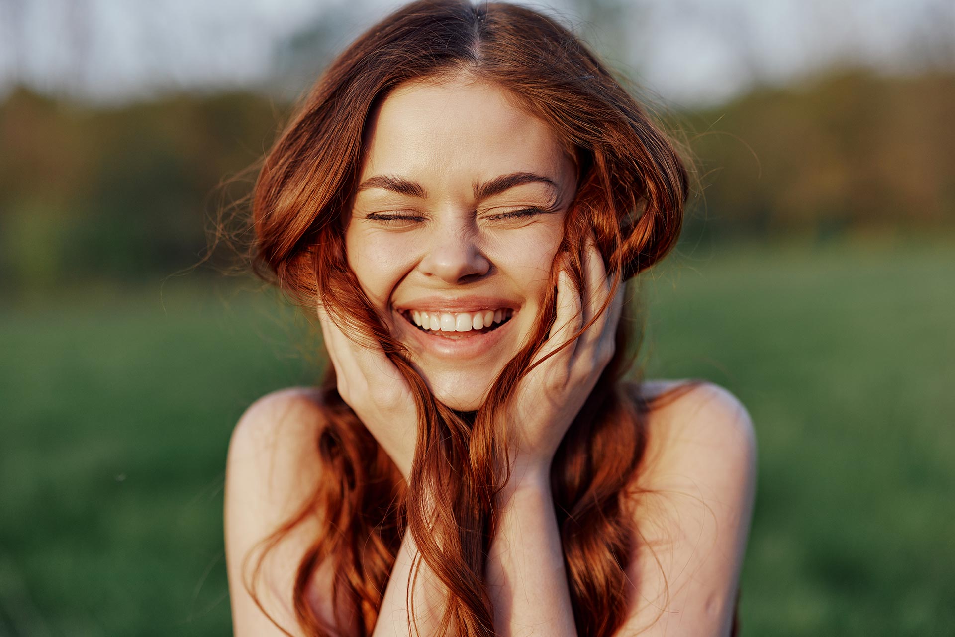 A young woman with long red hair is shown outdoors, smiling broadly, with her hands clasped near her face.