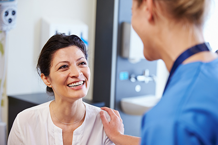 The image features two individuals in a medical setting a woman with dark hair wearing a white top, smiling at another person who appears to be a nurse or healthcare professional, standing next to her with a stethoscope around their neck and a name tag on their shirt, both are engaged in a friendly interaction.